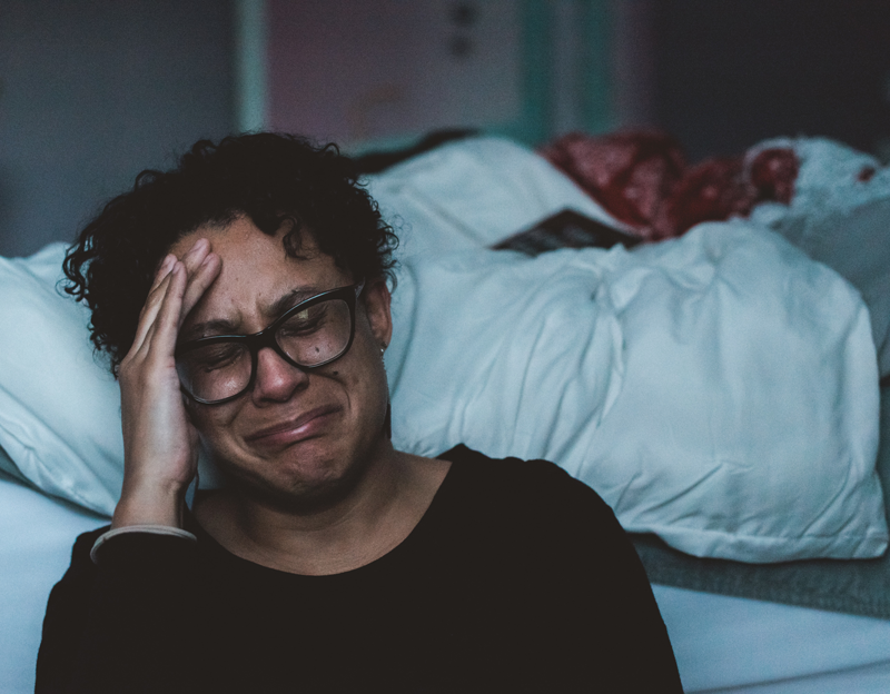 Depression/Anxiety/OCD brown skinned female with black short curly hair and black glasses crying on the floor next to a bed.